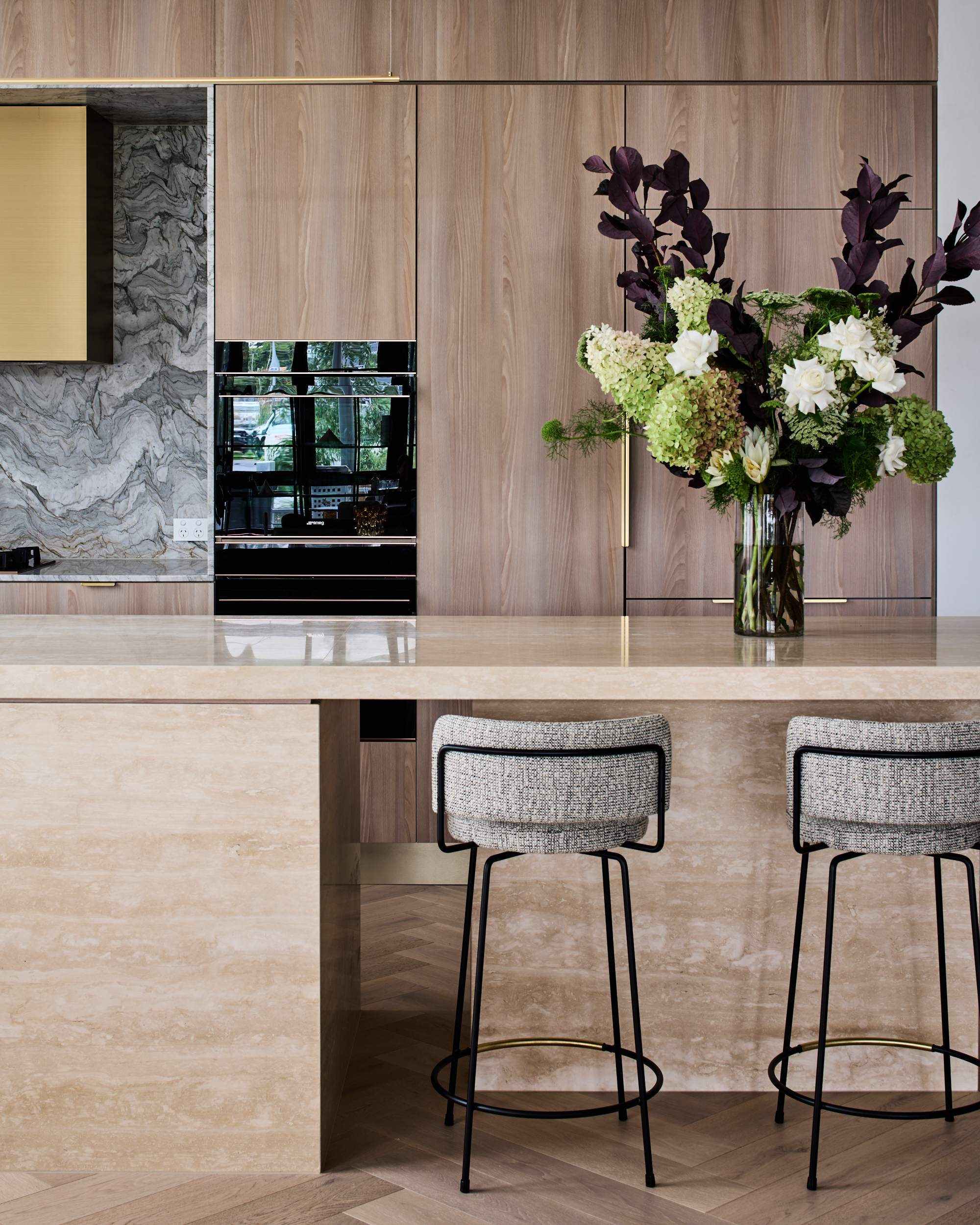 Luxury kitchen with travertine island bench, light timber joinery, stone splashback and a large floral display